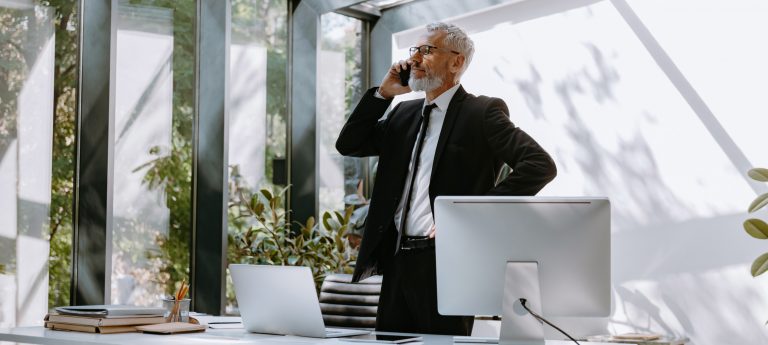 Confident mature businessman talking on mobile phone while standing near his working place in office
